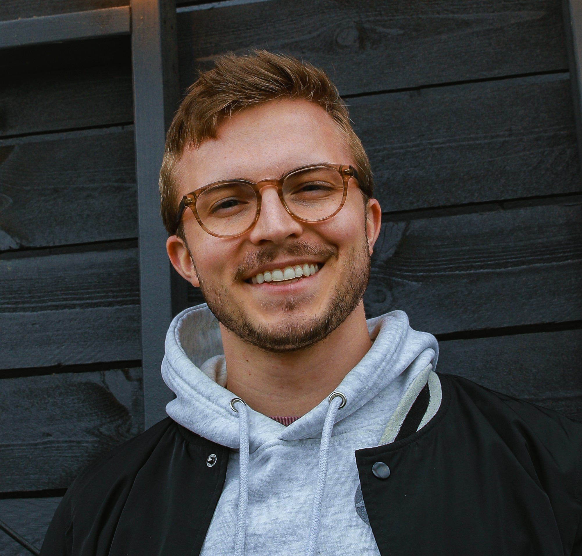 Smiling young man with glasses, hoodie, and jacket against a dark background.